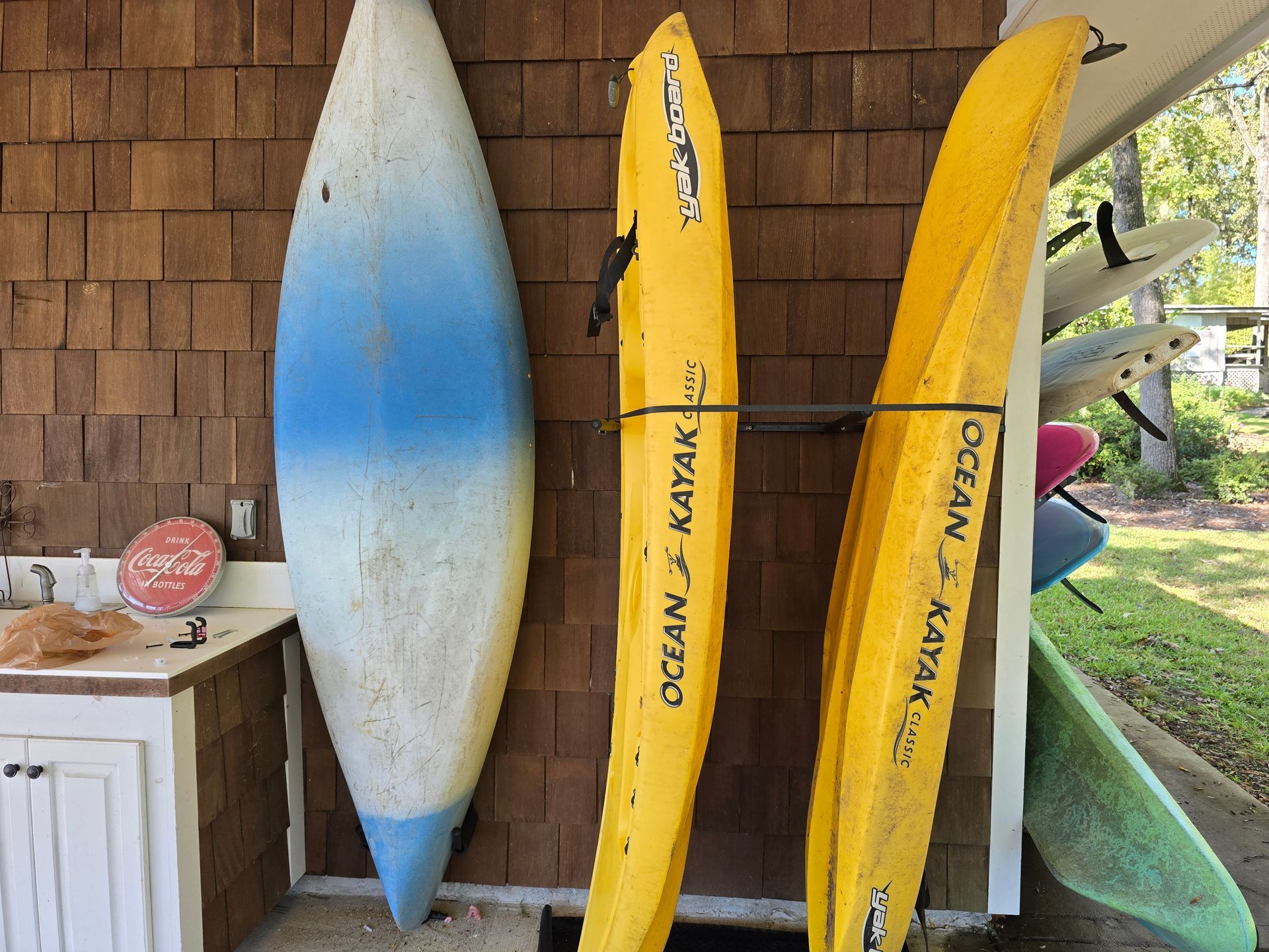 Three kayaks, two yellow and one blue, leaned against a wooden wall near an outdoor area.