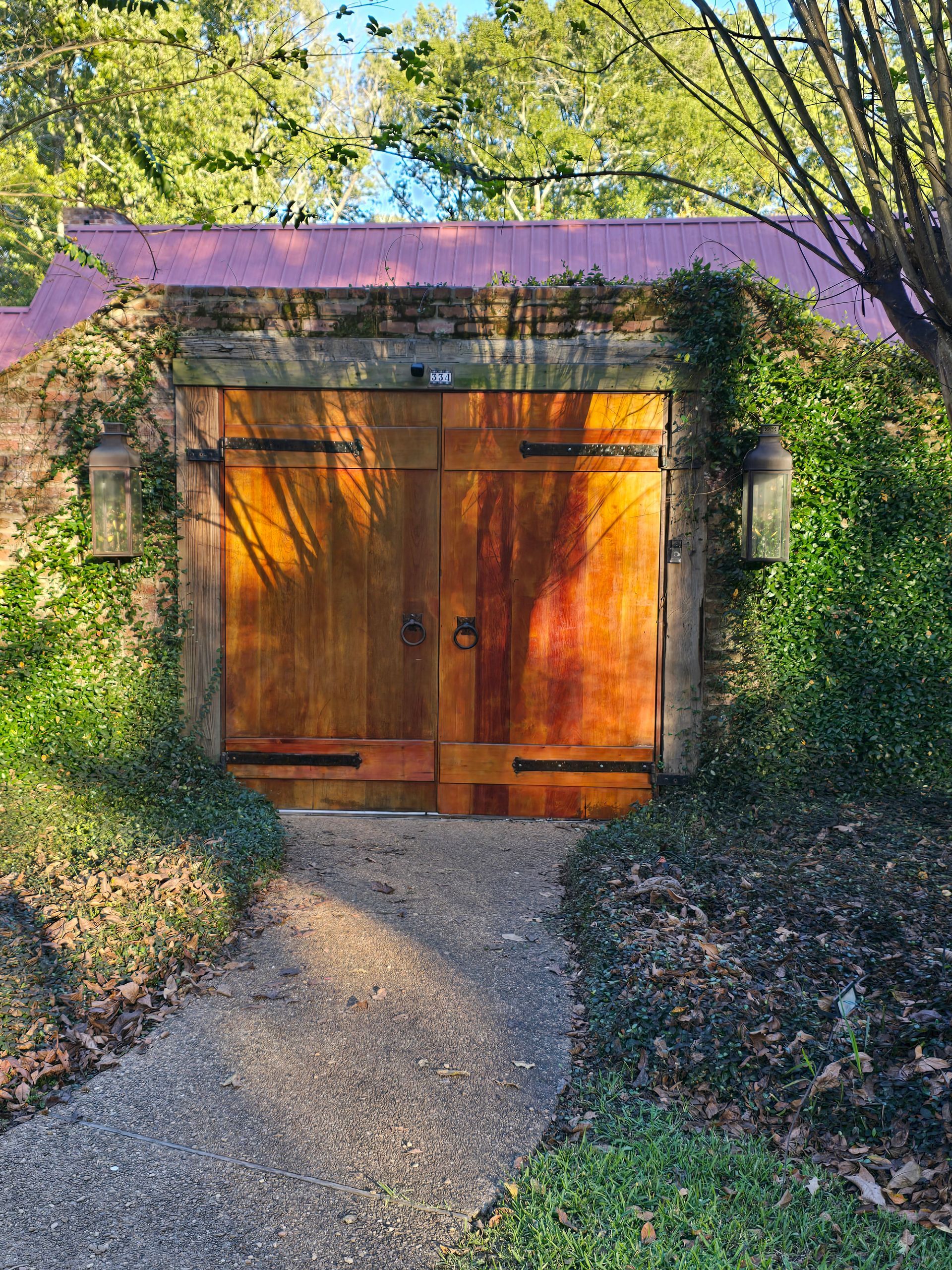 Wooden double doors in a brick wall, surrounded by greenery. Pathway leads to the entrance.