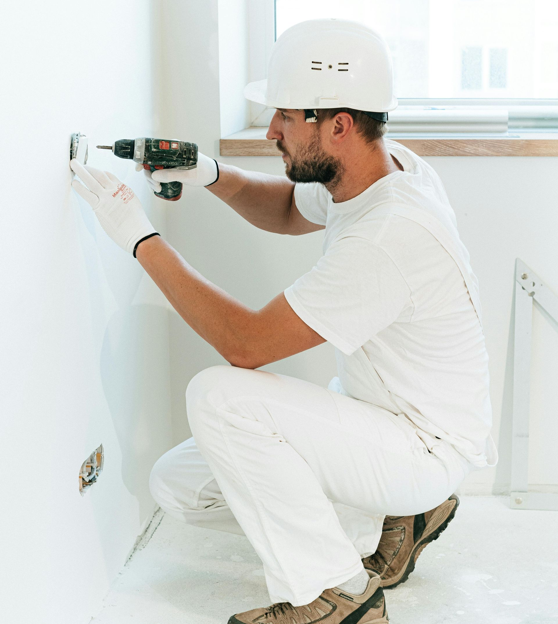 Construction worker in white overalls and hardhat using a drill on a wall.