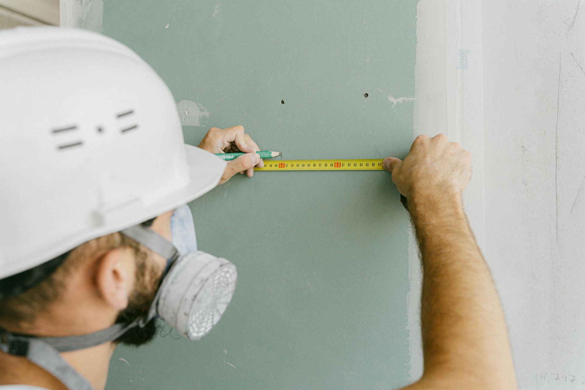 Construction worker measuring wall with tape measure, wearing hard hat and respirator.