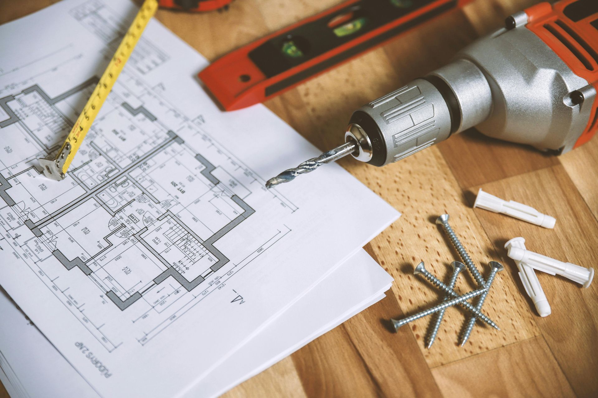 Blueprint, power drill, measuring tape, and hardware on a wooden surface, indicating construction work.