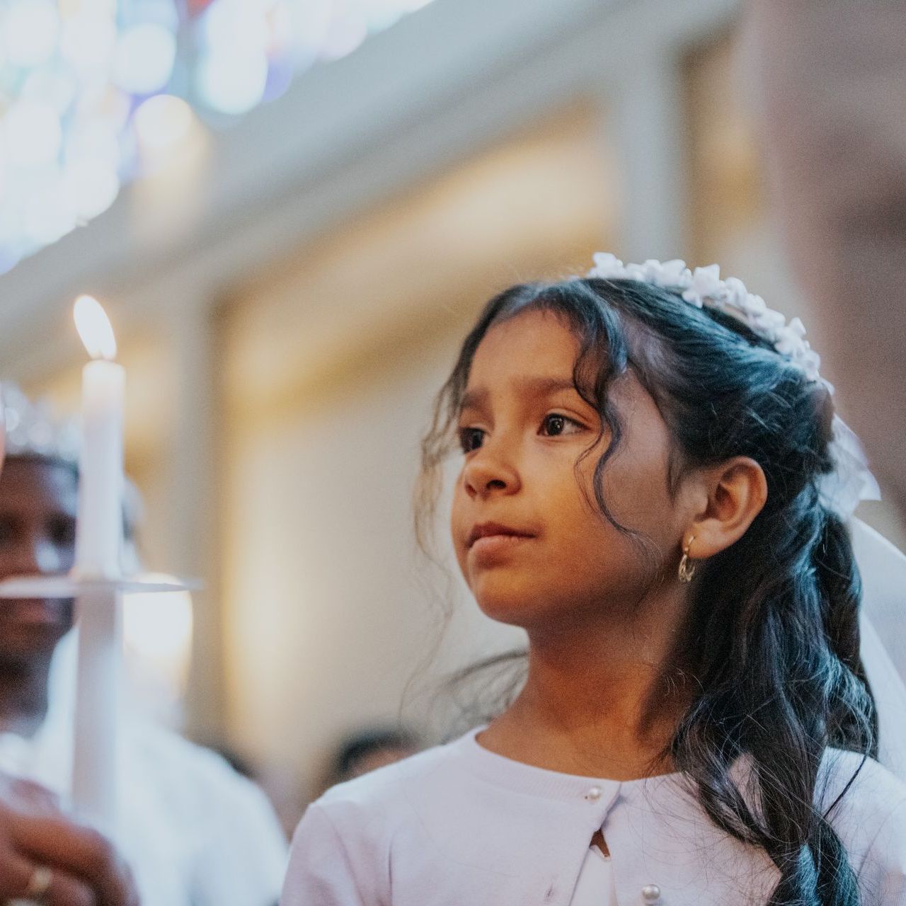 Young girl in a white dress and floral headband, looking ahead in a softly lit indoor ceremony