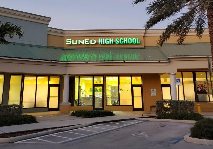 Suned High School entrance at dusk, with glowing yellow lights and palm trees outside.