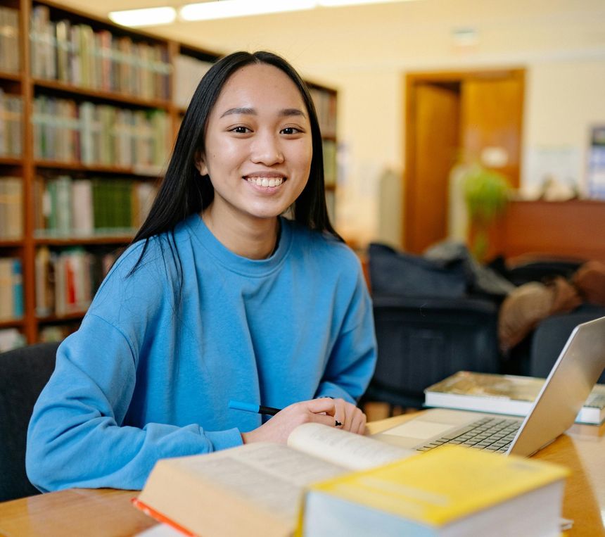 Student smiling at a library table with books and a laptop in a study area.