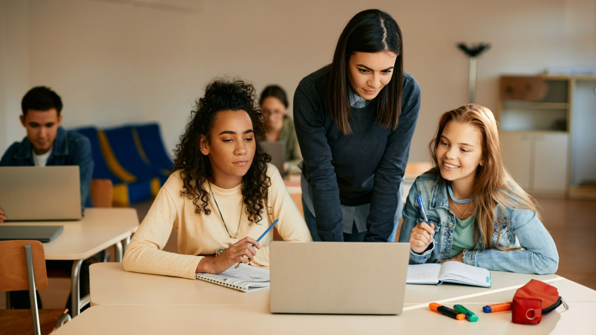 Three people gathered around a laptop at a table in a bright classroom, working together.