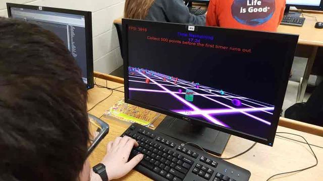 Person using a computer showing a neon 3D game scene on a monitor in a classroom setting