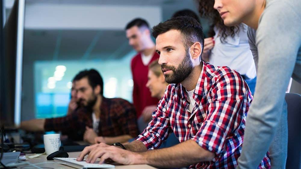 Team of coworkers collaborating at computers in a modern office, with one man focused on typing in the foreground