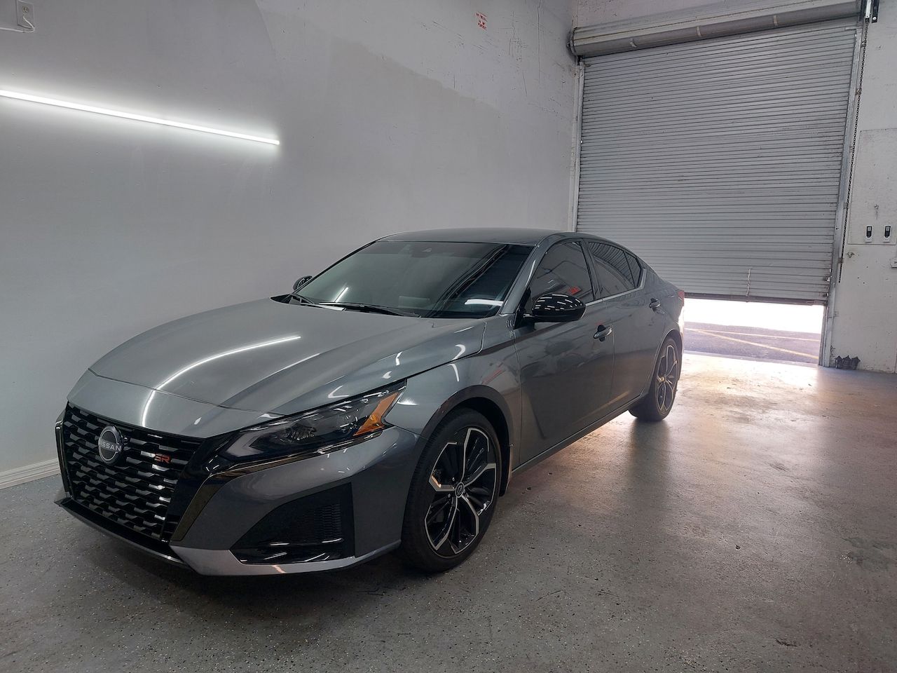A gray nissan altima is parked in a garage with a garage door open.