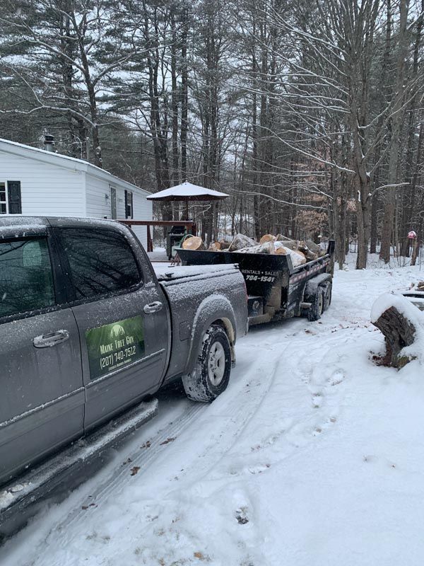 A gray truck towing a trailer loaded with wood in a snowy residential area.