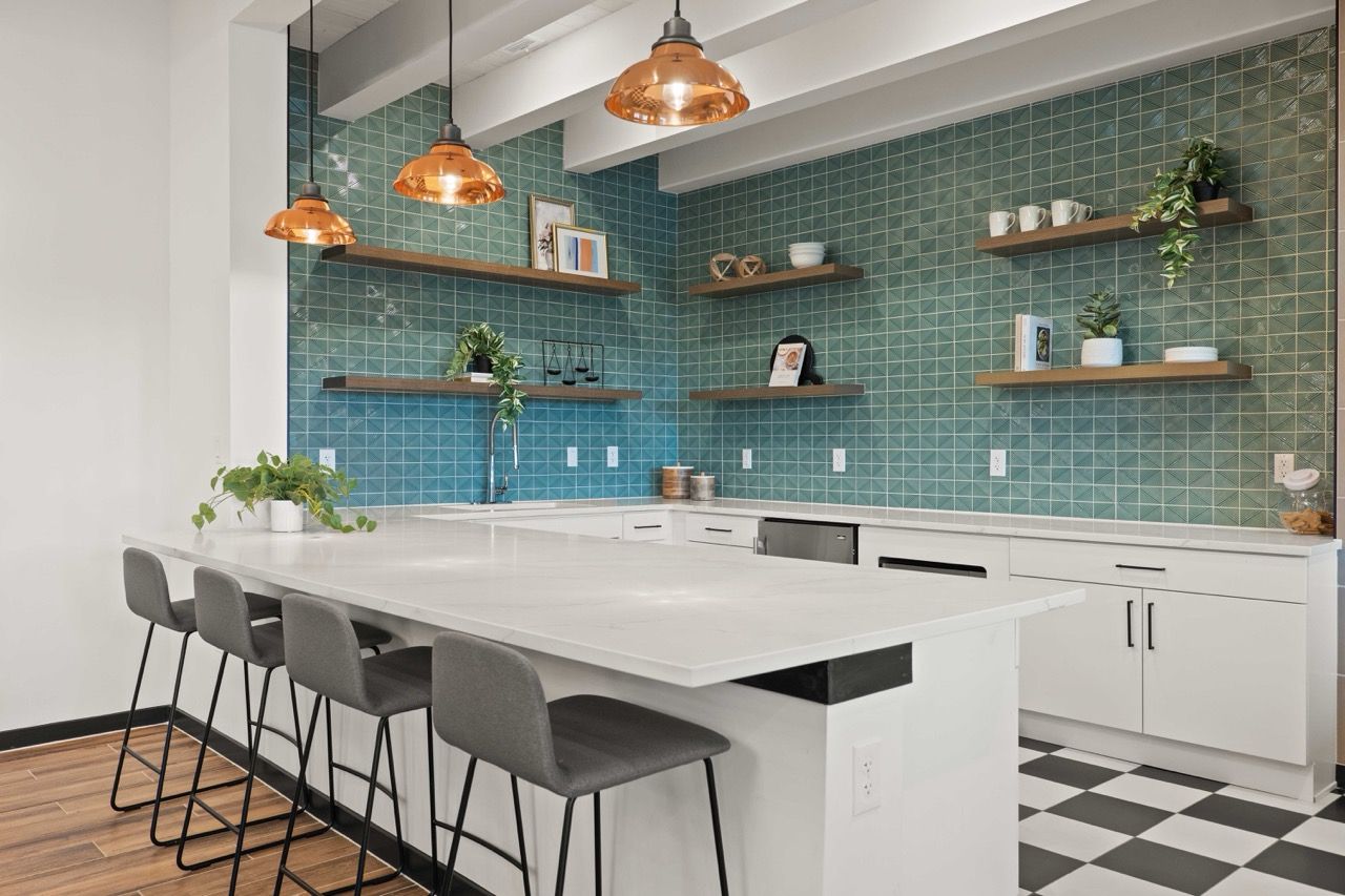Community kitchen with white cabinets, teal tile backsplash, an island and four gray stools.