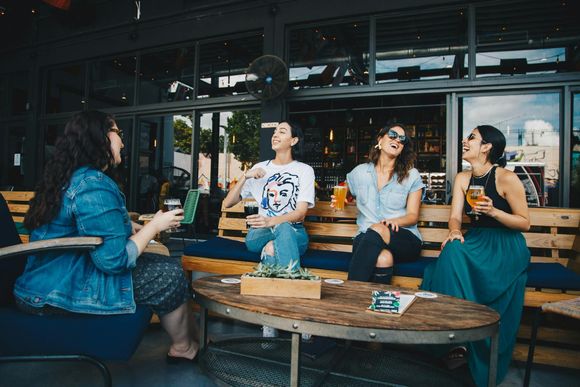 Group of friends sitting outside a caf&eacute; or brewery patio, laughing and chatting while holding drinks around a wooden table.