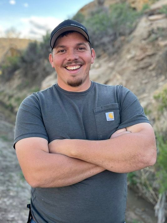 Smiling man in a gray T-shirt and black cap with arms crossed outdoors near a rocky hillside