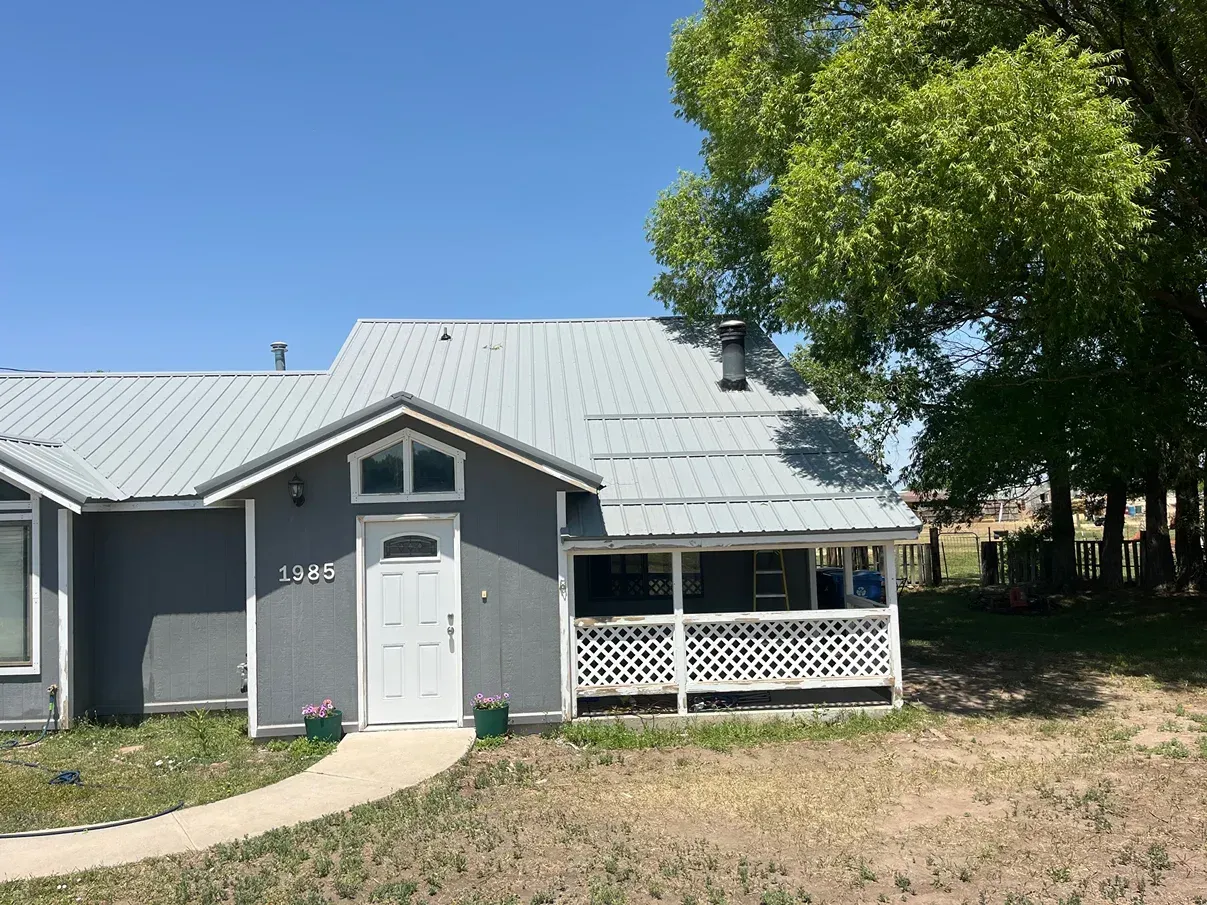 Gray house with white door and porch, metal roof. Green tree on right, clear blue sky.