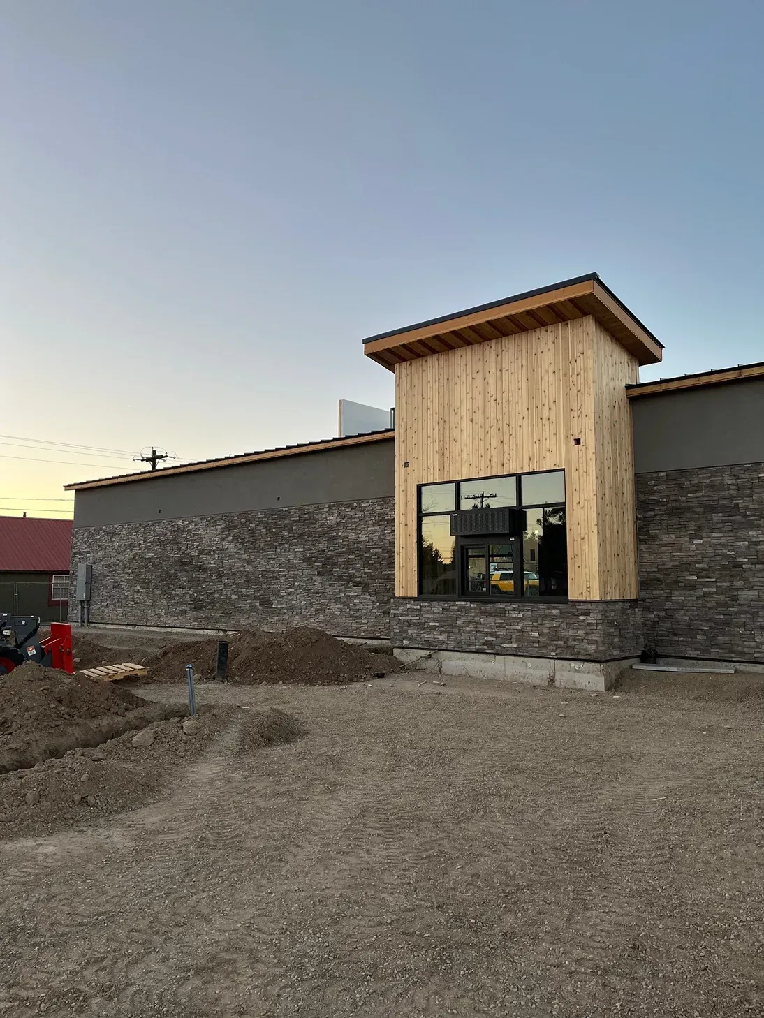 Building under construction; exterior with stone and wood accents. Gravel lot, window, and blue sky.
