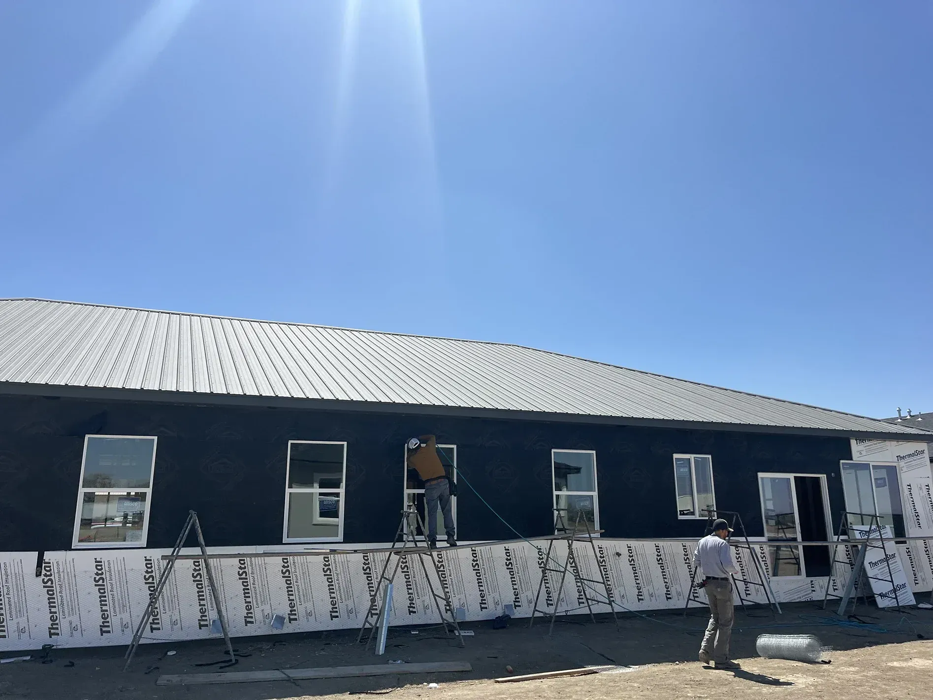 Construction workers installing windows on a long, dark building with a metal roof on a sunny day.