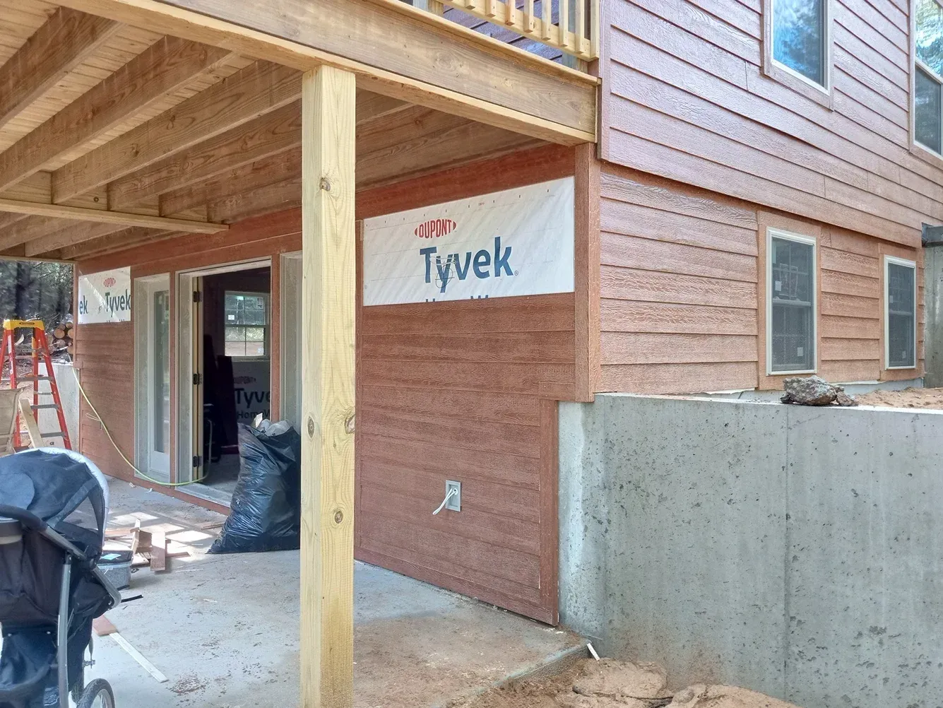 House under construction with brown siding, Tyvek wrap, and concrete foundation. Deck overhead.
