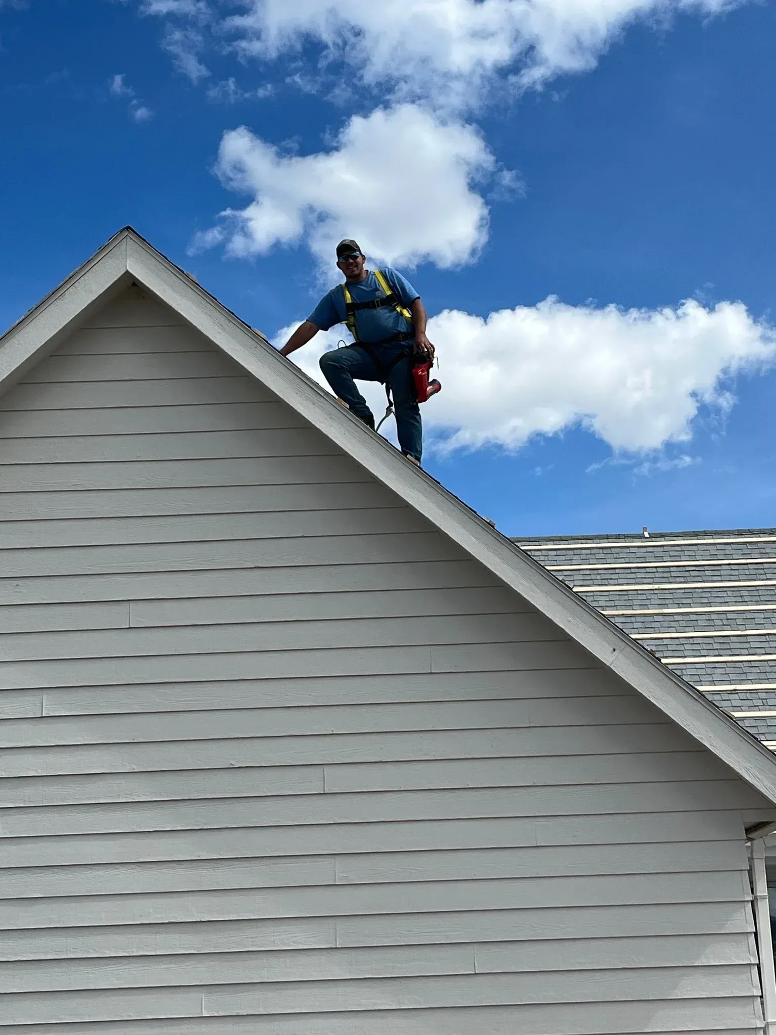 Person on a roof with a harness, checking shingles on a sunny day with clouds.