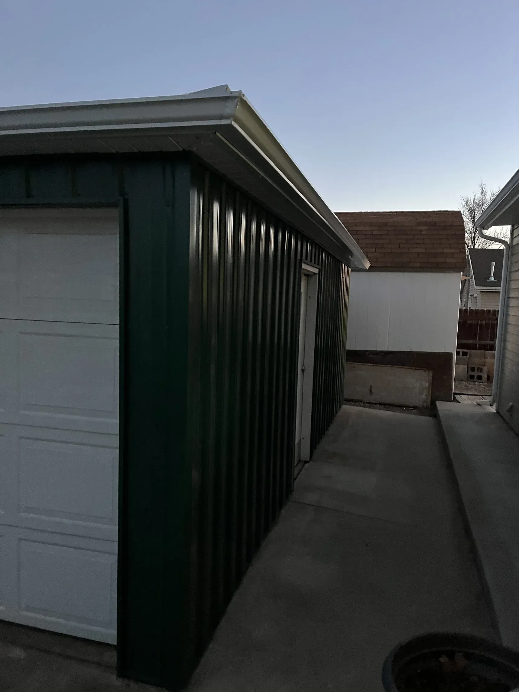 Green corrugated-metal sided garage with white door, concrete path, and nearby buildings.