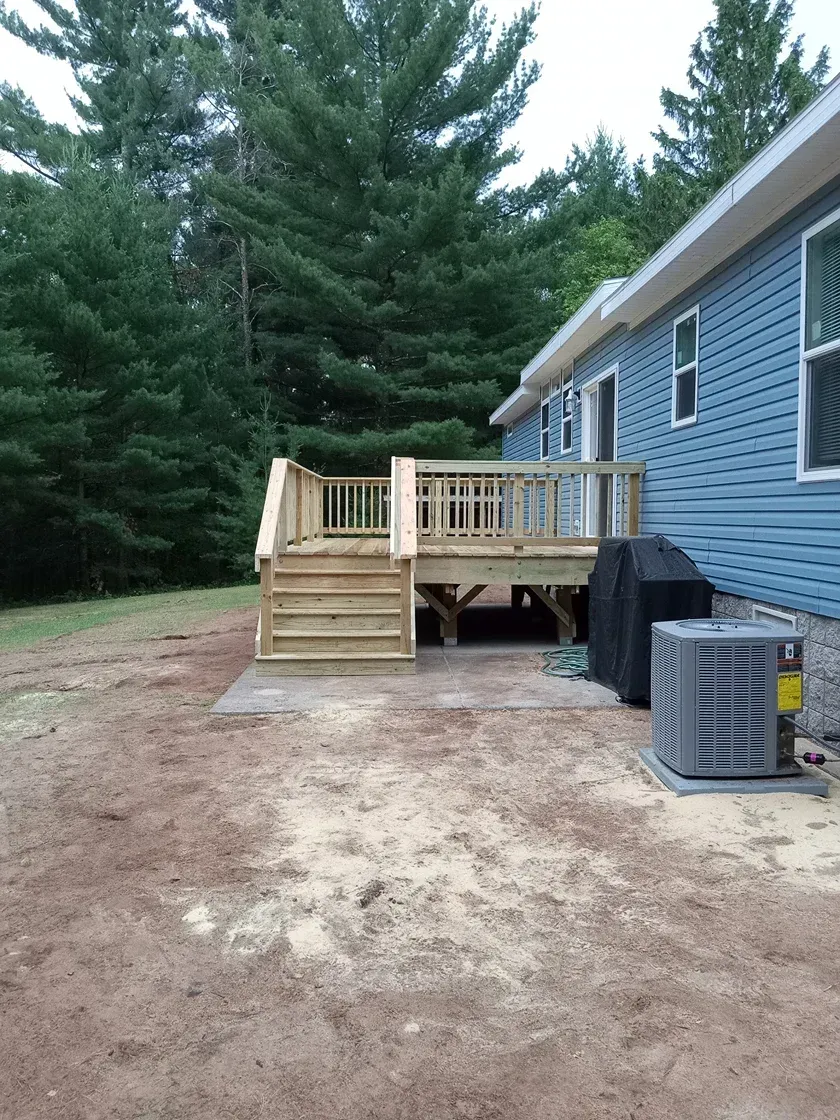 Wooden deck with stairs attached to a blue-sided house, surrounded by trees and dirt.