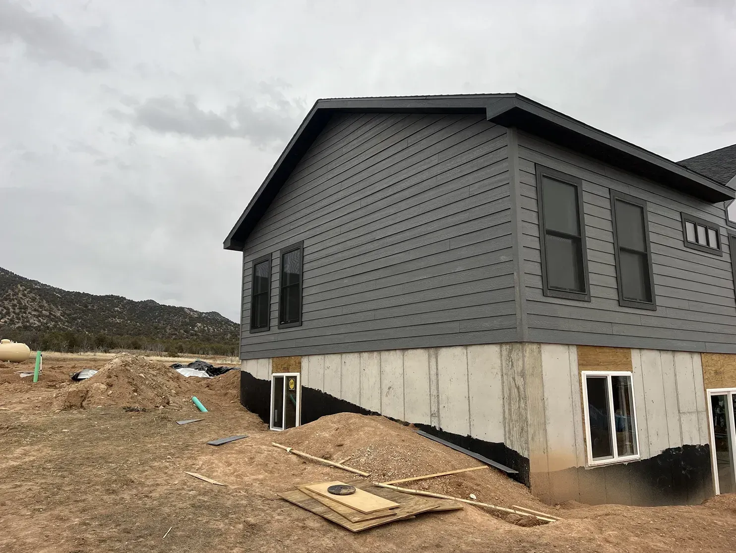 Two-story house under construction with gray siding and concrete foundation on a dirt lot under a cloudy sky.