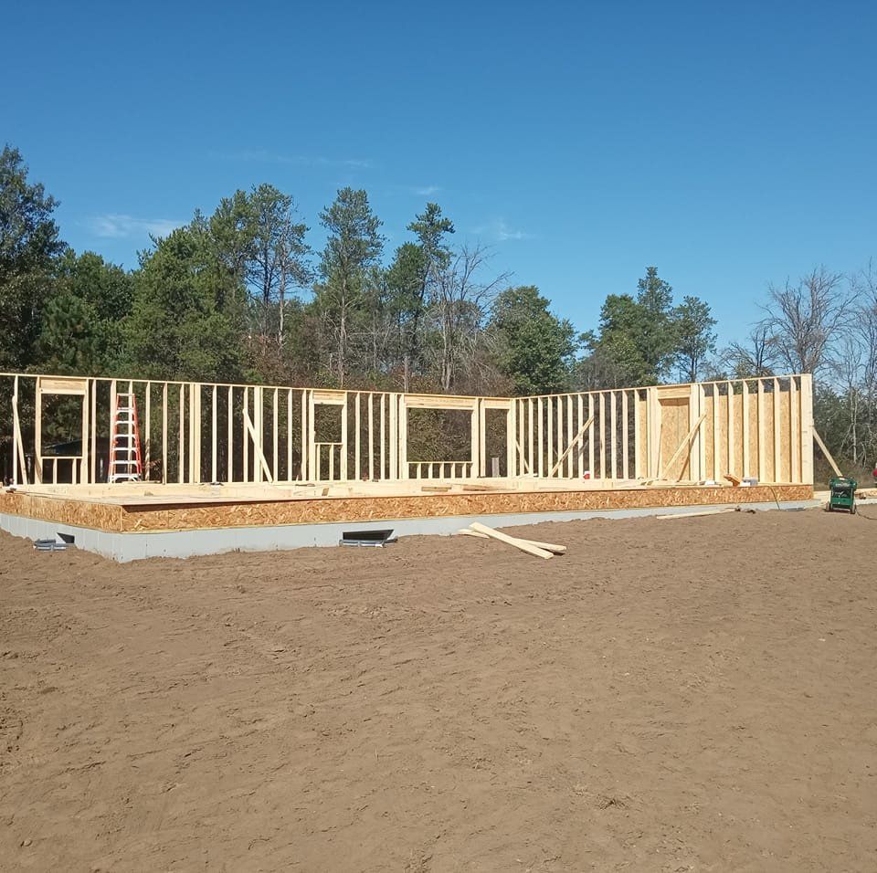 Wooden framing of a house under construction on a concrete foundation, with a blue sky background and trees.