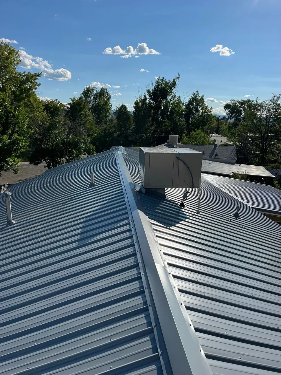 Metal roof with coiled pipes and rooftop ventilation against a blue sky with trees.