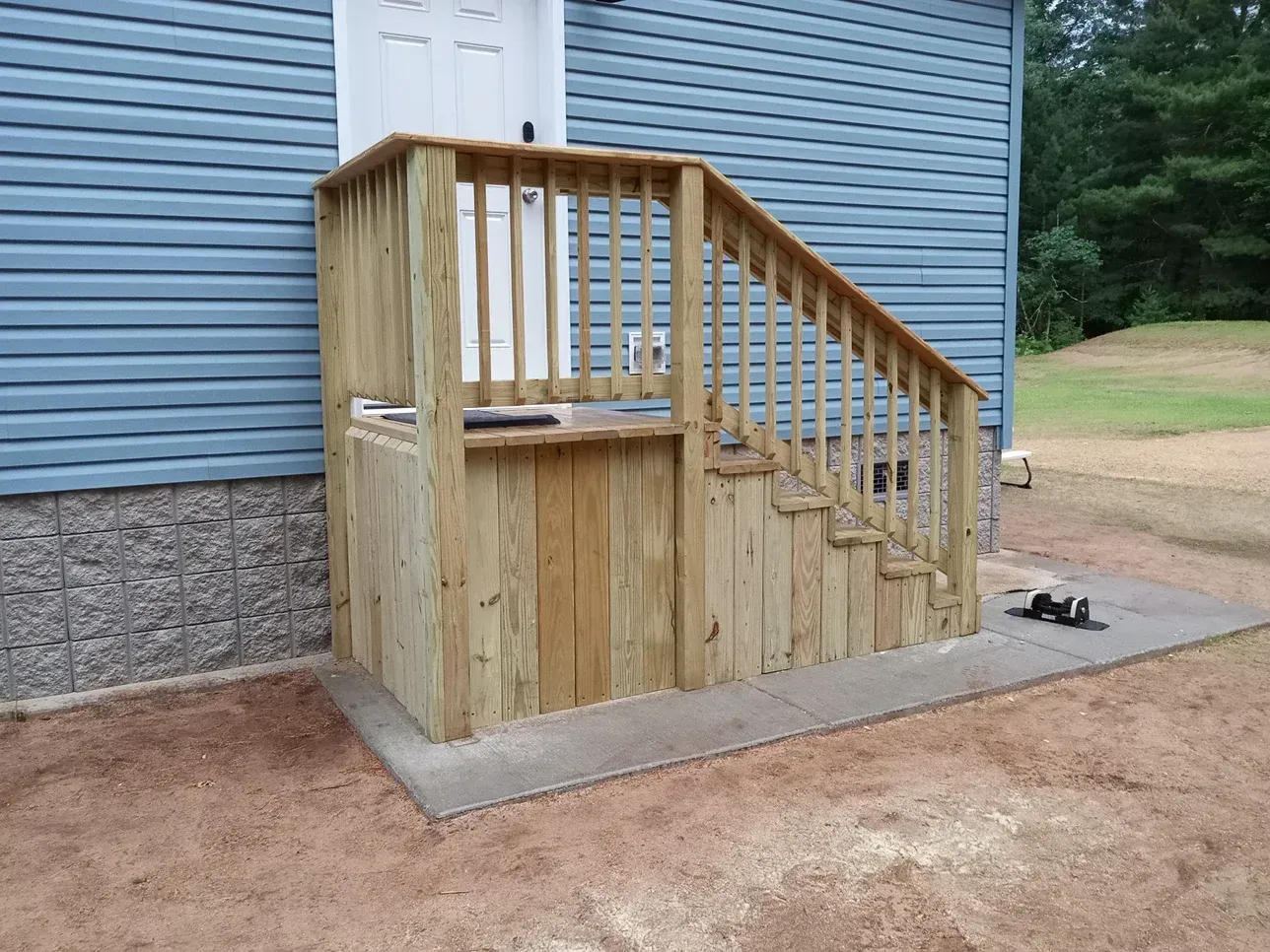 Wooden staircase and landing with railing, leading to a white door on a blue-sided house.