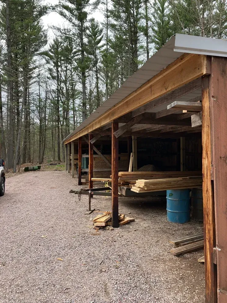 Lumber storage shed with wood stacked inside, gravel drive, and tall pine trees in the background.