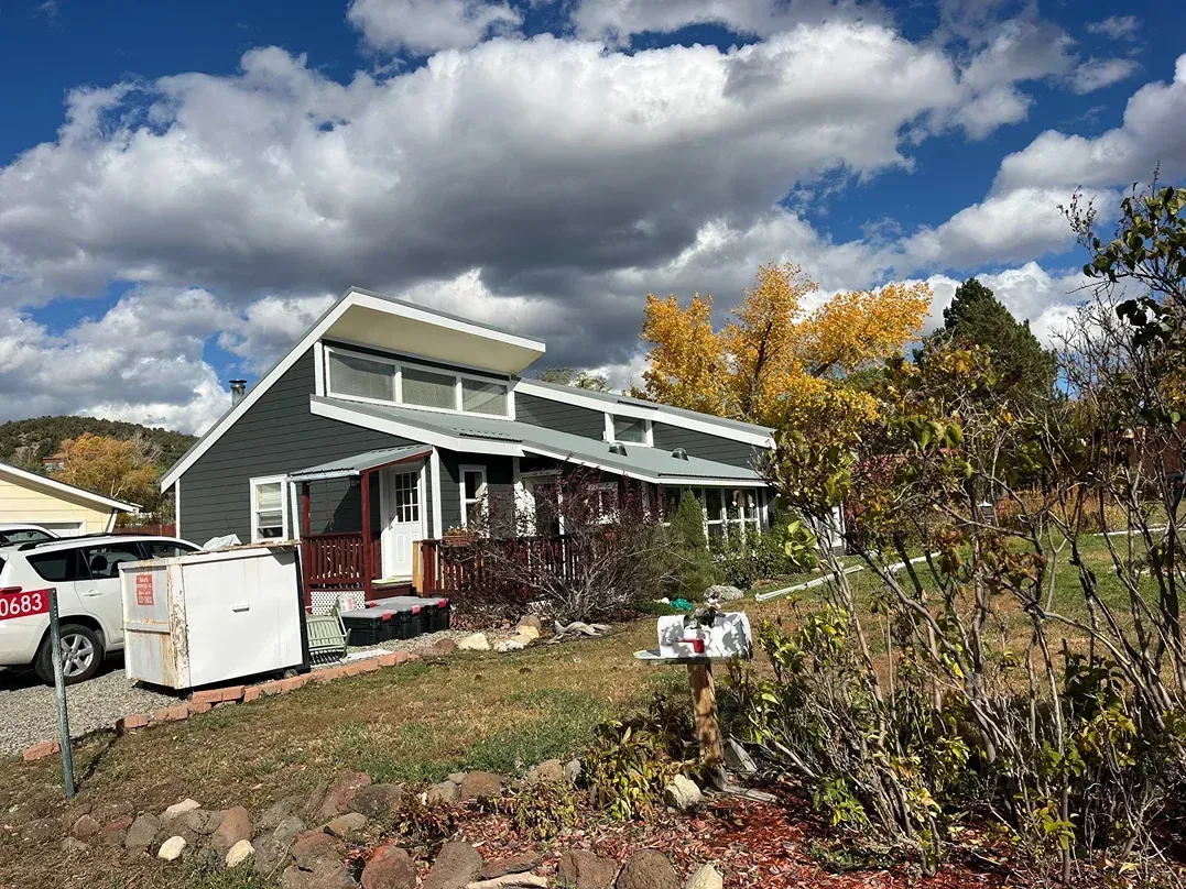 Gray house with white trim, porch, and a slanted roof under a cloudy blue sky.