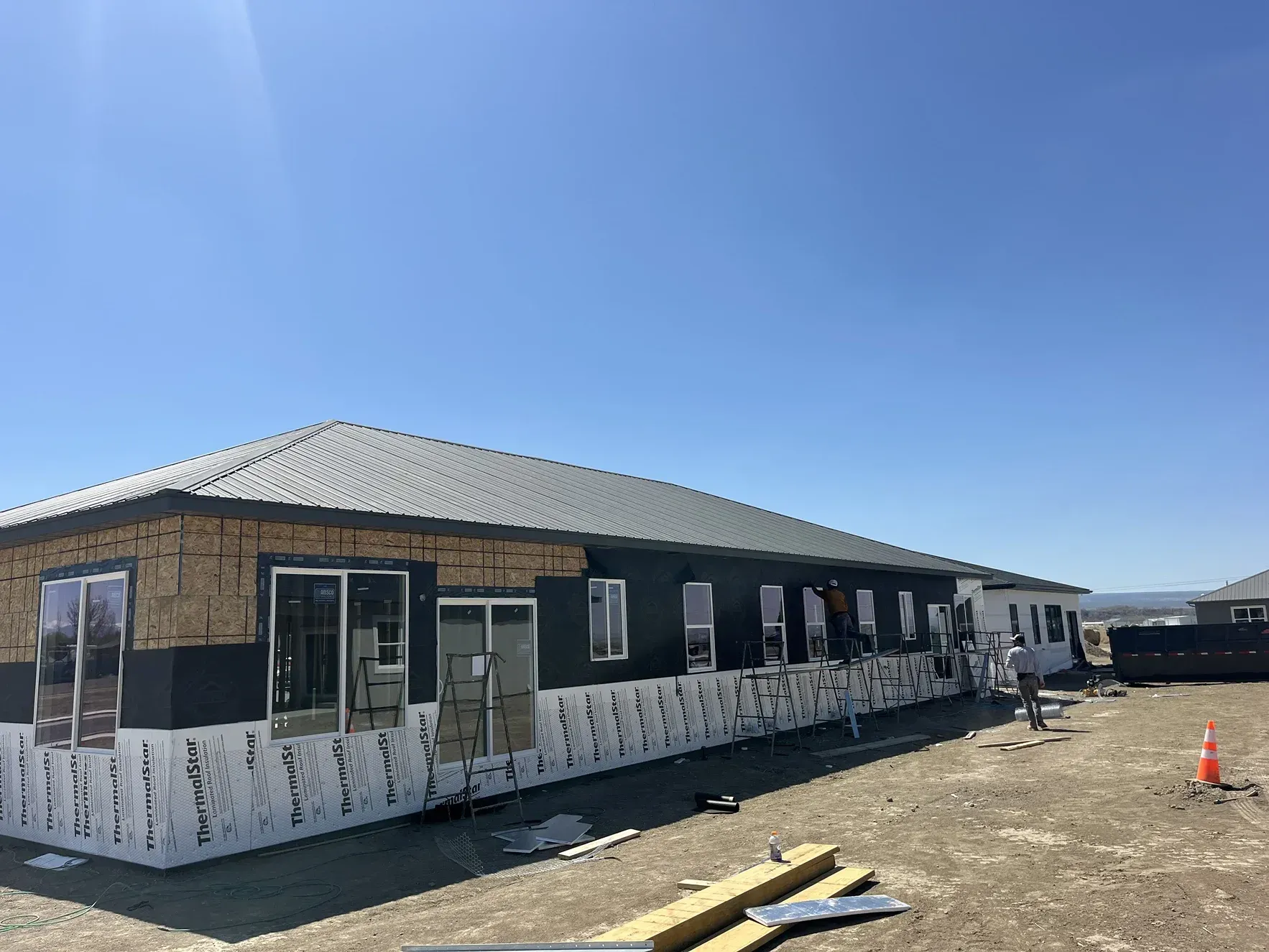 Construction site: single-story building with roof, windows, exterior walls in progress under a clear blue sky.