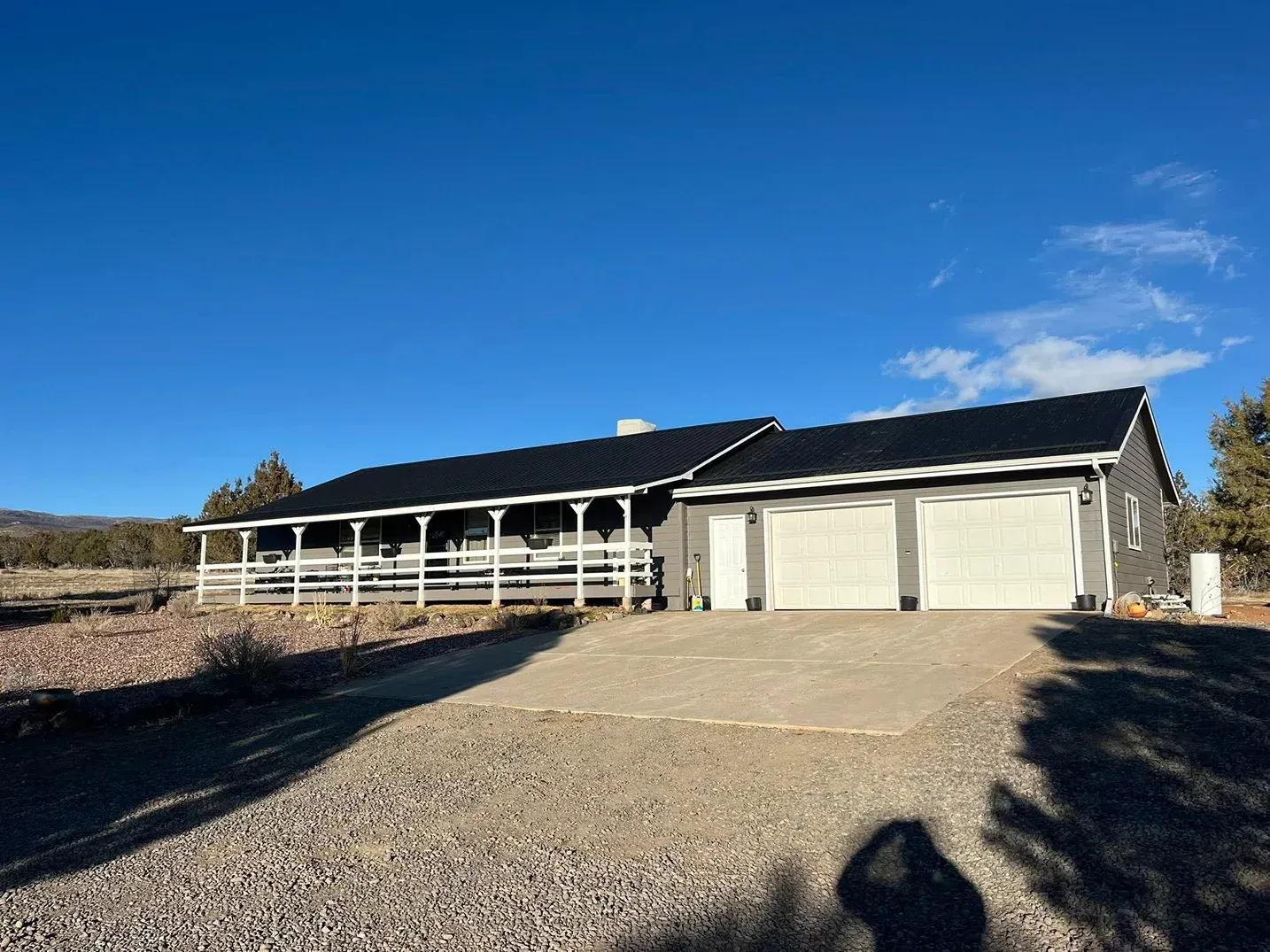 Gray ranch-style house with black roof, covered porch, and two-car garage under a blue sky.