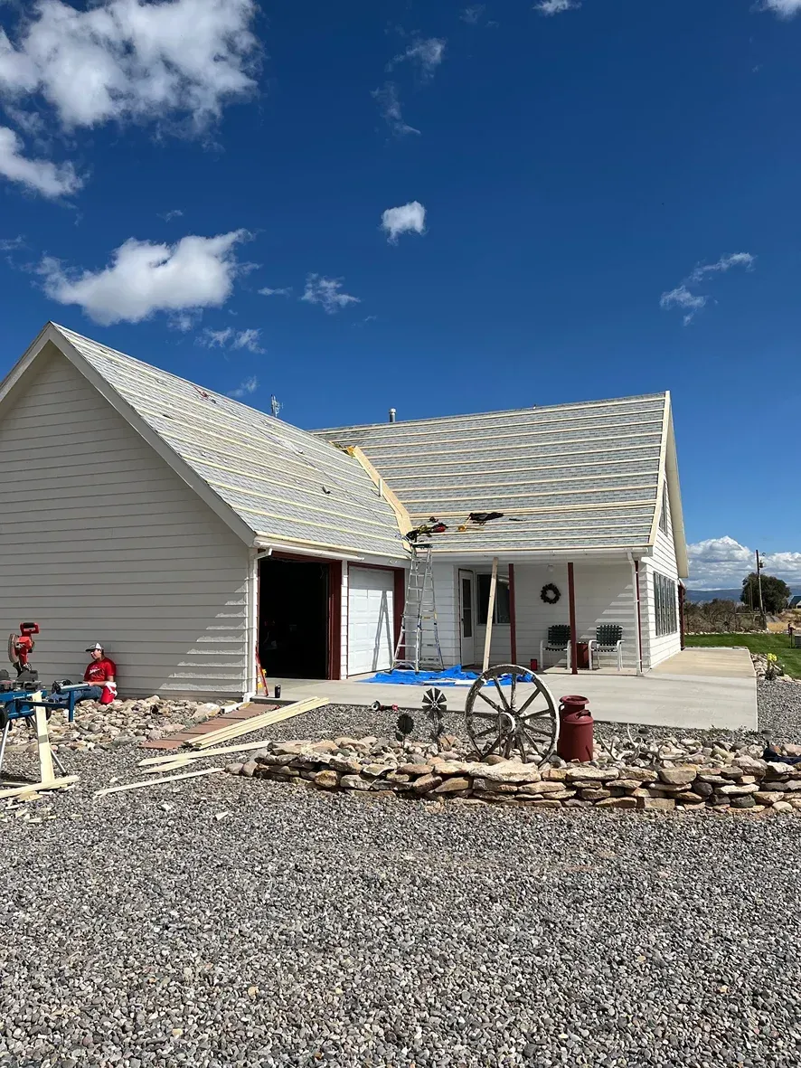 White house with a gray shingle roof under a blue sky, gravel yard, and porch.