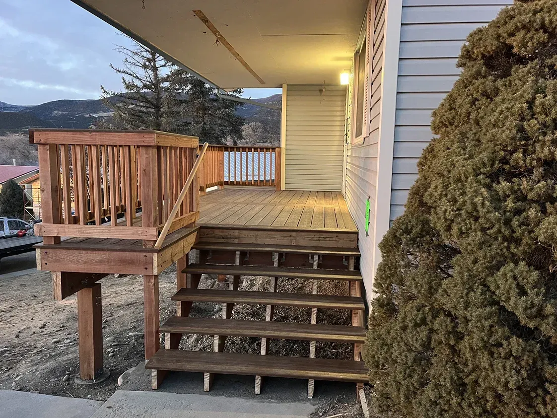 Wooden deck with steps leading to a house. Brown railing, stairs, and deck. House is light blue with green accents.