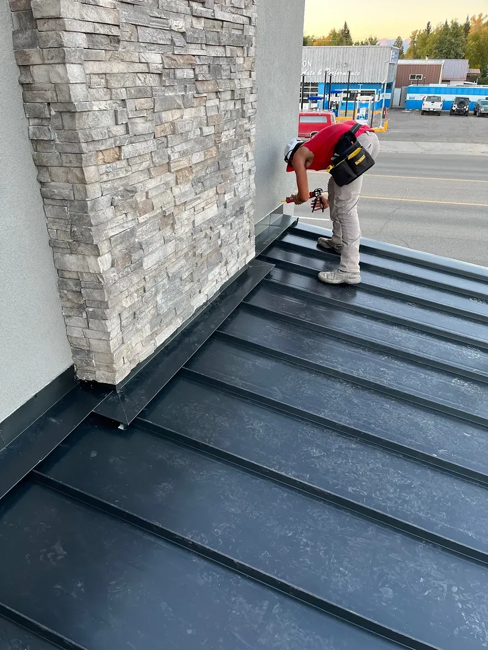 Roofer in red shirt working on a dark metal roof next to a stone chimney.