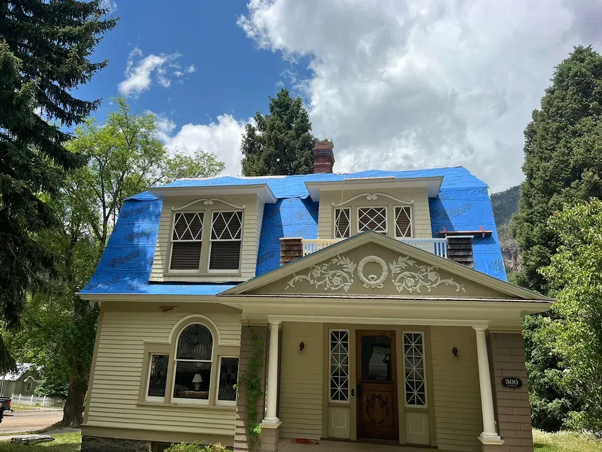 A two-story house with a blue tarp roof, surrounded by trees under a partly cloudy sky.