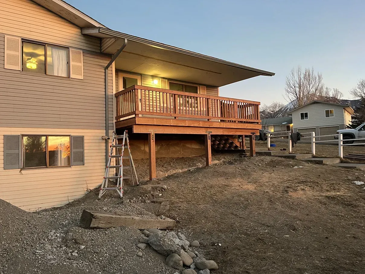 A house with a wooden deck; a ladder leans against the deck. The ground is covered in gravel.