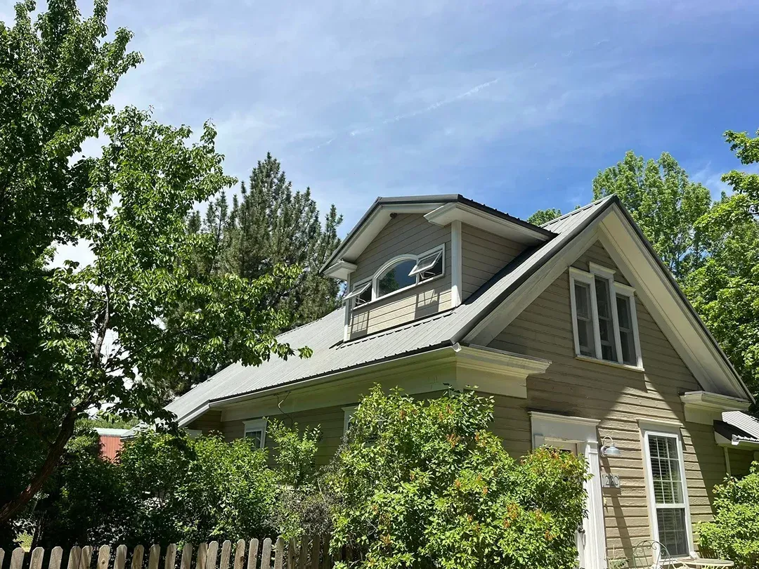 Tan house with gray roof, surrounded by green trees under a blue sky.