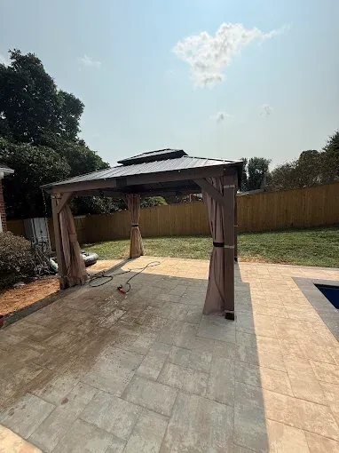 Gazebo on a stone patio with tan curtains, a grassy yard, and a blue sky.