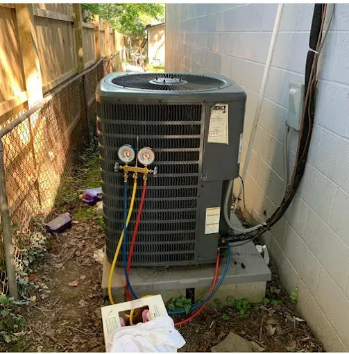 AC unit being serviced outdoors, gauges attached. Yellow, red, and blue hoses connected. Unit is grey.