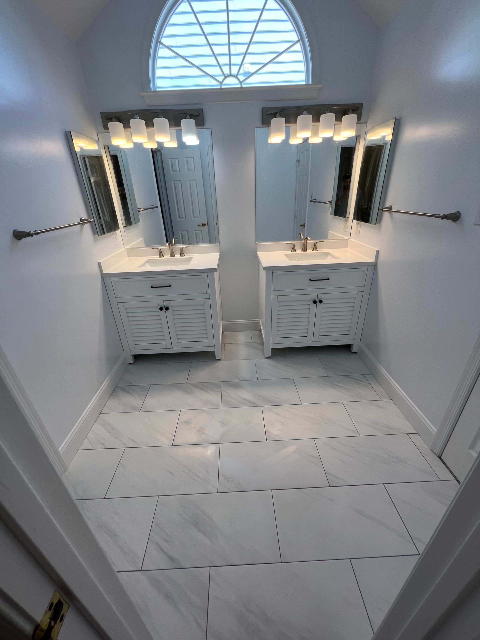 Bathroom with two white vanities, mirrors, lights, and tiled floor. Arched window above.