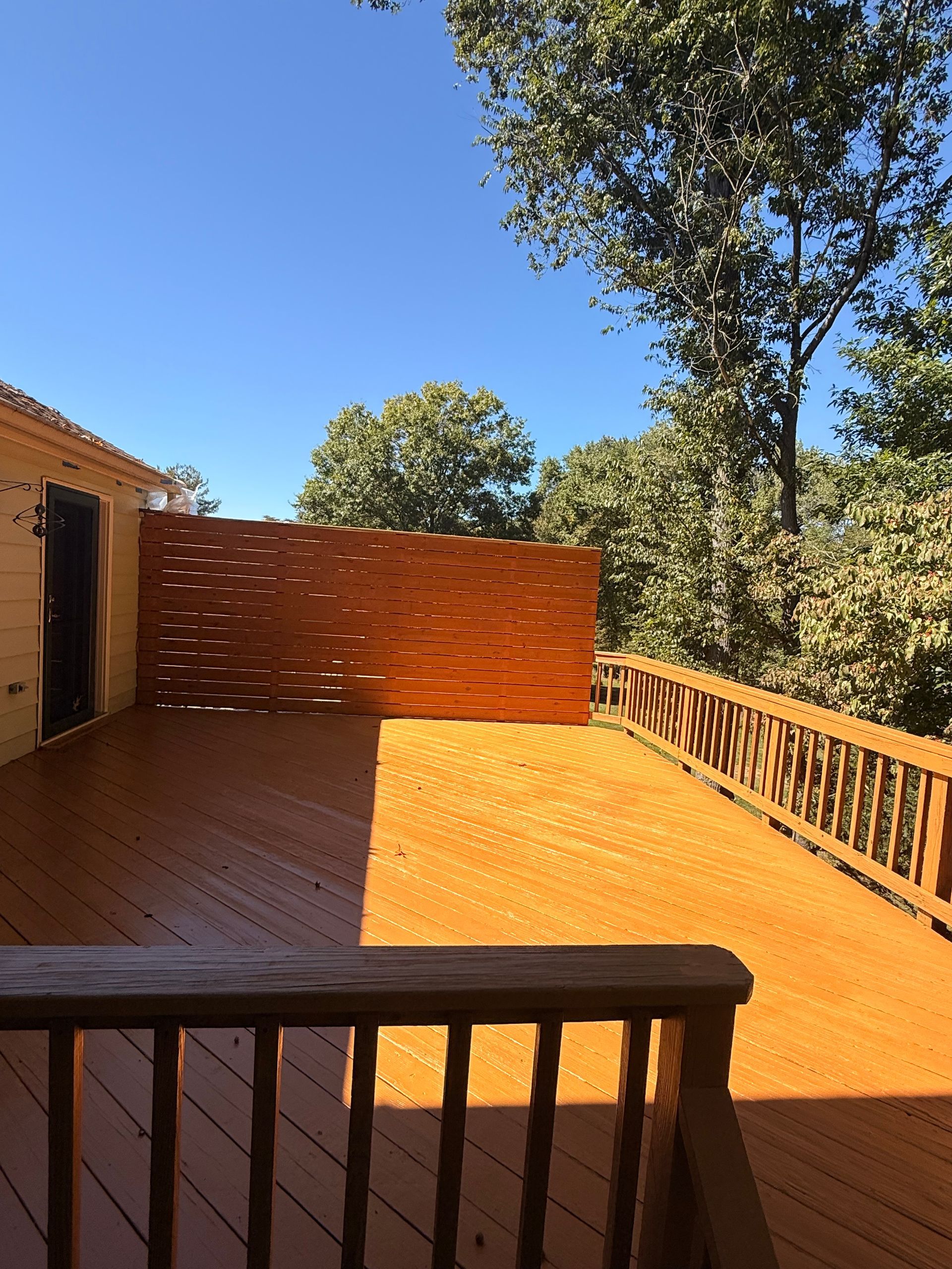 Wooden deck with a stained orange finish, partial privacy screen, and railing. Sunny day, trees in background.