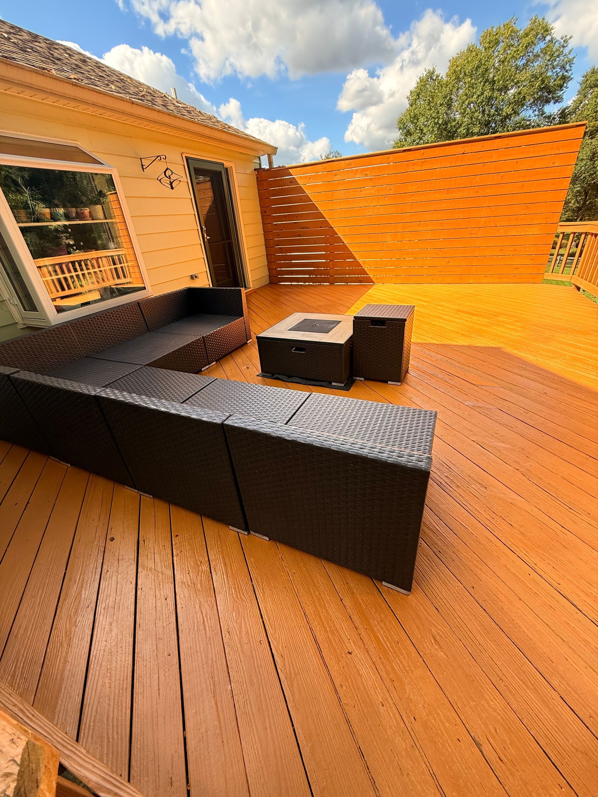 Outdoor deck with brown wooden boards, black sectional seating, and a fire pit. Sunny day, blue sky.