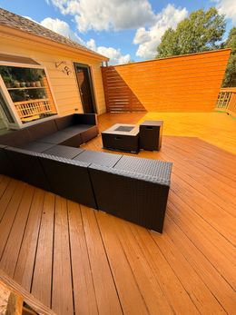 Outdoor deck with brown wooden boards, black sectional seating, and a fire pit. Sunny day, blue sky.