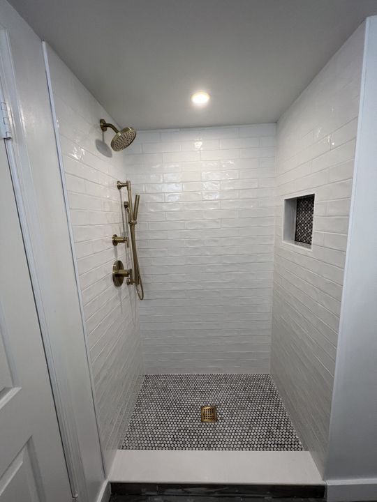 White tiled shower with bronze fixtures and black and white patterned floor.