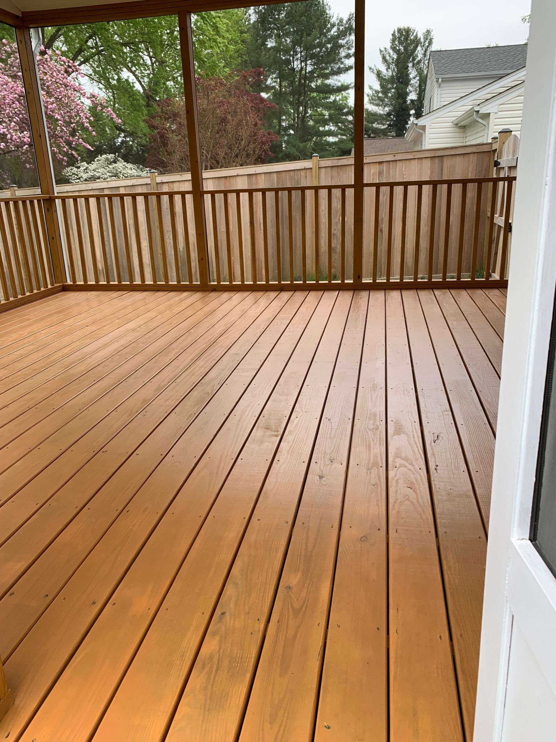 Wooden deck with railings, enclosed by screen and fence. Trees and house in background.