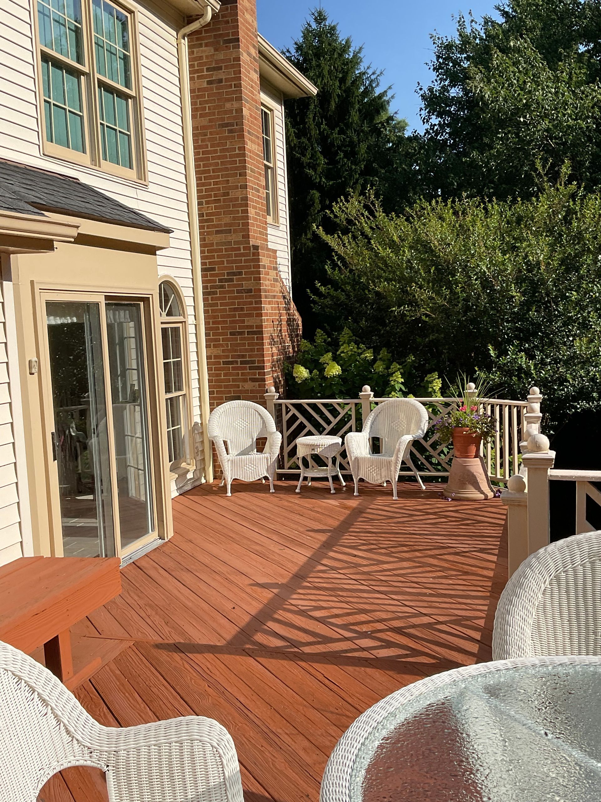 Wooden deck with white wicker furniture, red brick chimney, and lush greenery.