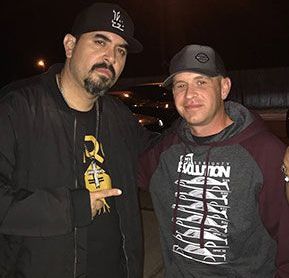 Two men are posing for a picture together in a parking lot.