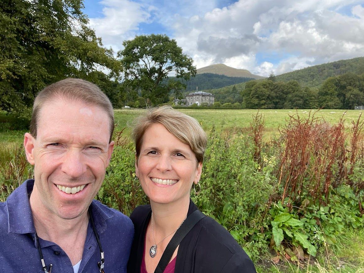 Couple smiles for selfie in field with a castle, mountains and trees in background.