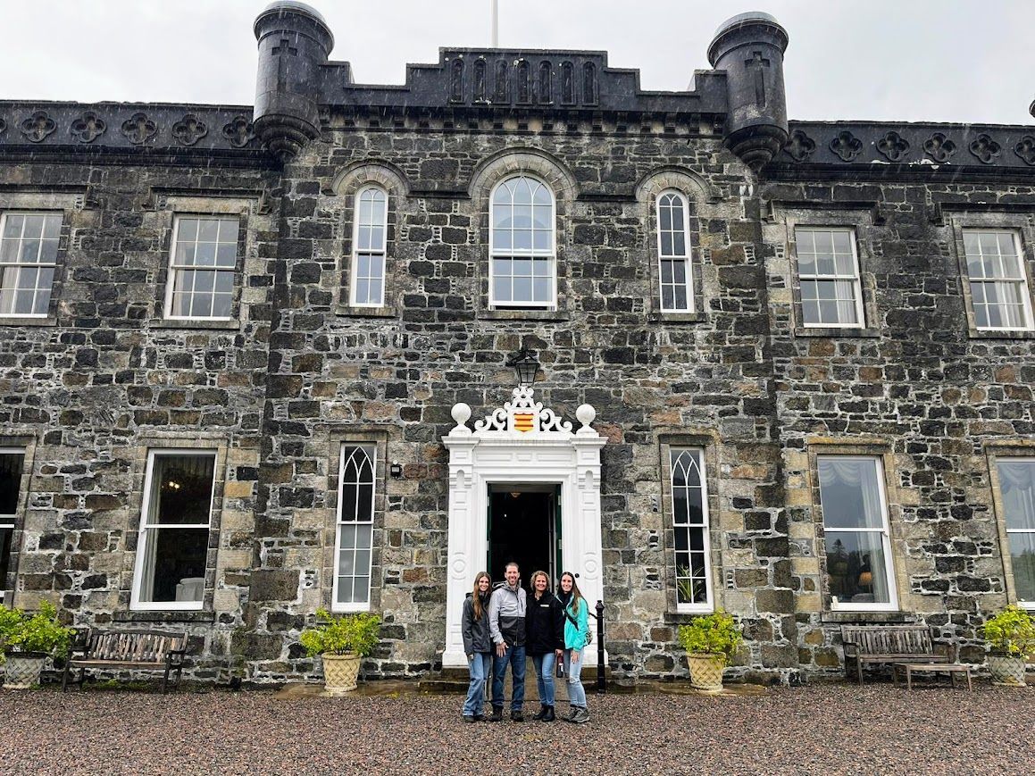 Group of people pose in front of stone castle with arched doorway and windows. Gray cloudy sky overhead.