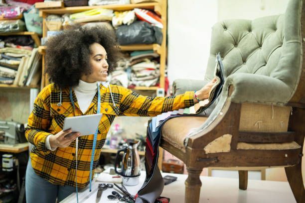 Upholsterer reupholstering chair in workshop with tablet, fabric samples, and tools around.